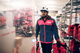 Lächelnder Flink Lieferant steht nachts mit seinem E-Bike und pinker Liefertasche vor einem beleuchteten Gebäude. 