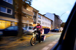Lächelnder Flink Lieferant steht nachts mit seinem E-Bike und pinker Liefertasche vor einem beleuchteten Gebäude. 