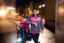 Lächelnder Flink Lieferant steht nachts mit seinem E-Bike und pinker Liefertasche vor einem beleuchteten Gebäude. 